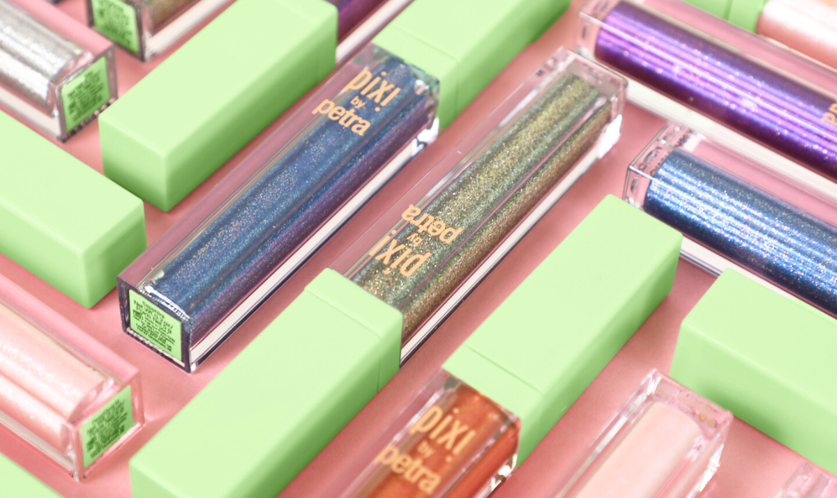 A close-up, angled view of Pixi by Petra liquid lip glosses in various shimmer shades, arranged diagonally on a pastel pink background. The clear tubes reveal glittery blue, gold, purple, and orange shades, while the matte light green caps are visible.