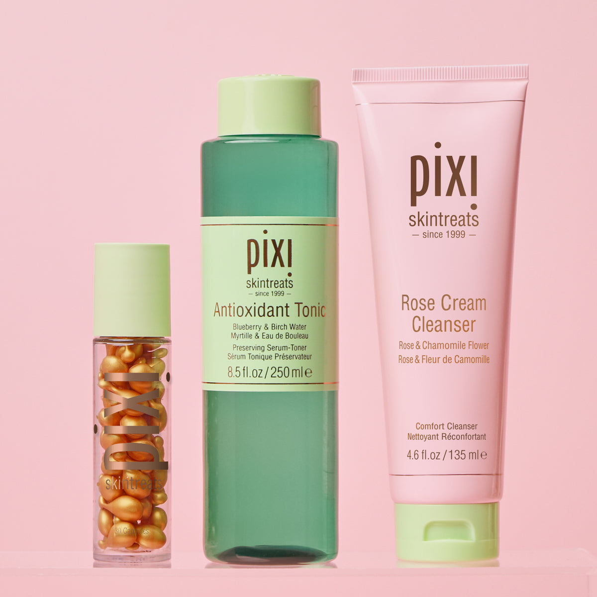 Three Pixi Skintreats skincare products arranged on a soft pink background: a small clear bottle with gold capsules, a translucent green bottle labeled Antioxidant Tonic, and a pink tube labeled Rose Cream Cleanser.