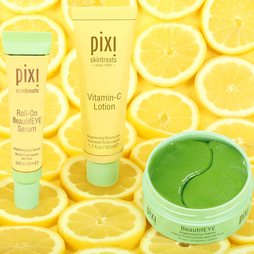 Three Pixi Skintreats BeautifEYE products arranged on a background of sliced lemons: a Roll-On BeautifEYE Serum tube, a Vitamin-C Lotion tube, and an open BeautifEYE jar containing bright green eye patches. The products use yellow and mint-green packaging. view 2